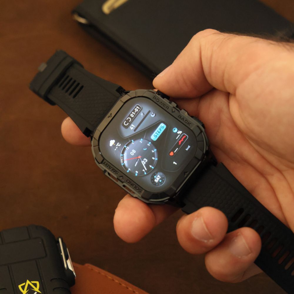 Person holding a black smartwatch with a digital display, showing time and various icons.